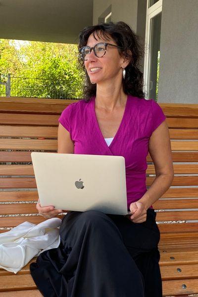 Claudia Potschigmann sitzt auf einer Bank, mit Macbook am Schoss und blickt in die Ferne
