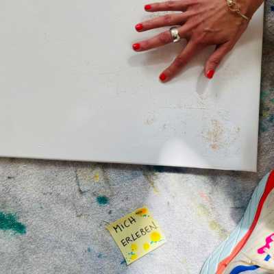 Claudias Hand ist hier zu sehen, auf eine weiße Leinwand tupfend, mit einem Post-It-Zettel am Boden, auf dem "Mich erleben" geschrieben steht.