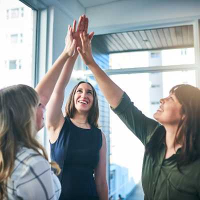 Drei junge Frauen feiern ihren Erfolg als Unternehmerin, indem sie high five mit den Händen zusammenschlagen, als Bild beim Artikel Zielstrebigkeit.